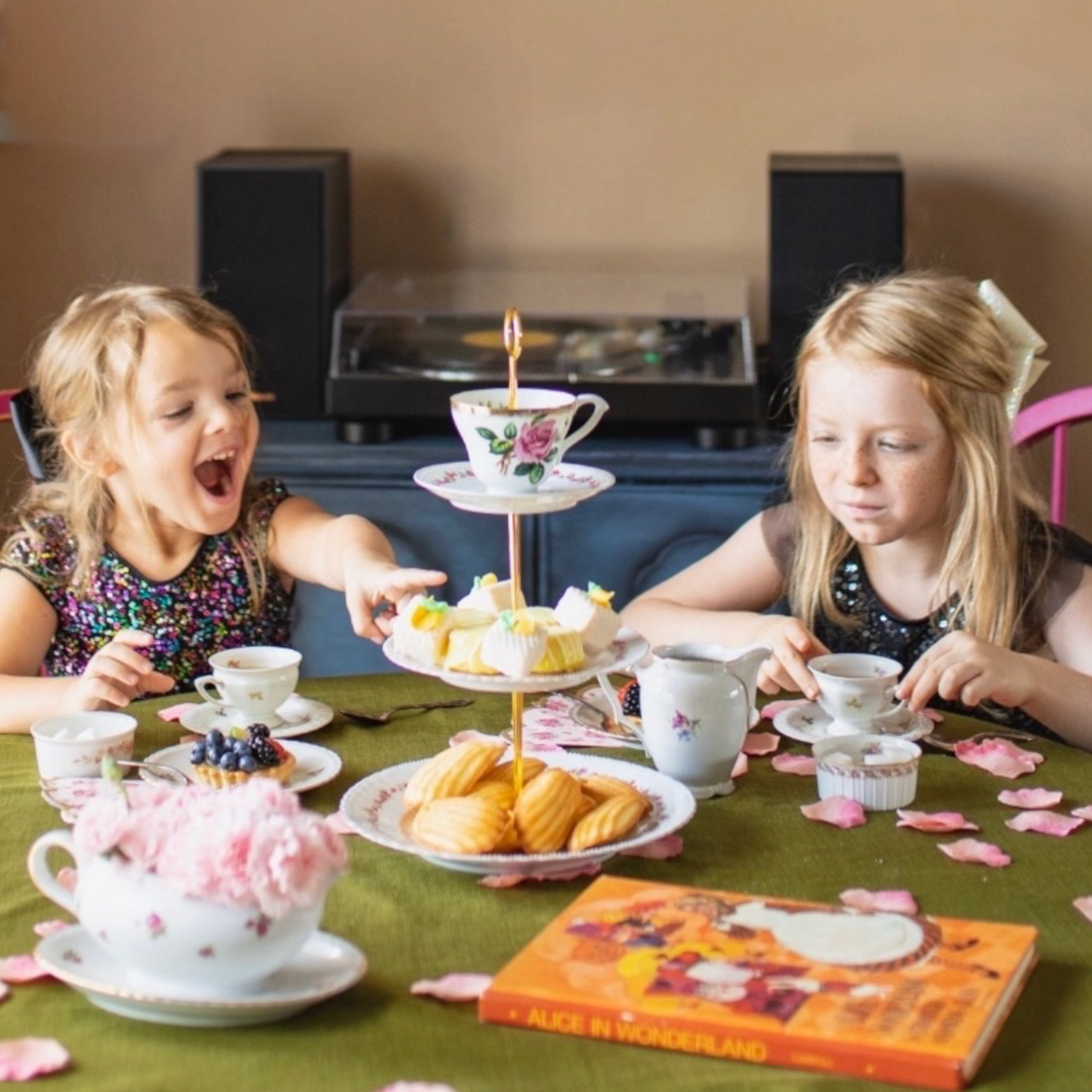 Two young girls enjoying a tea party on tiered tray from grandmother’s china. Eating pastries with Alice in Wonderland vibe in a cozy room.