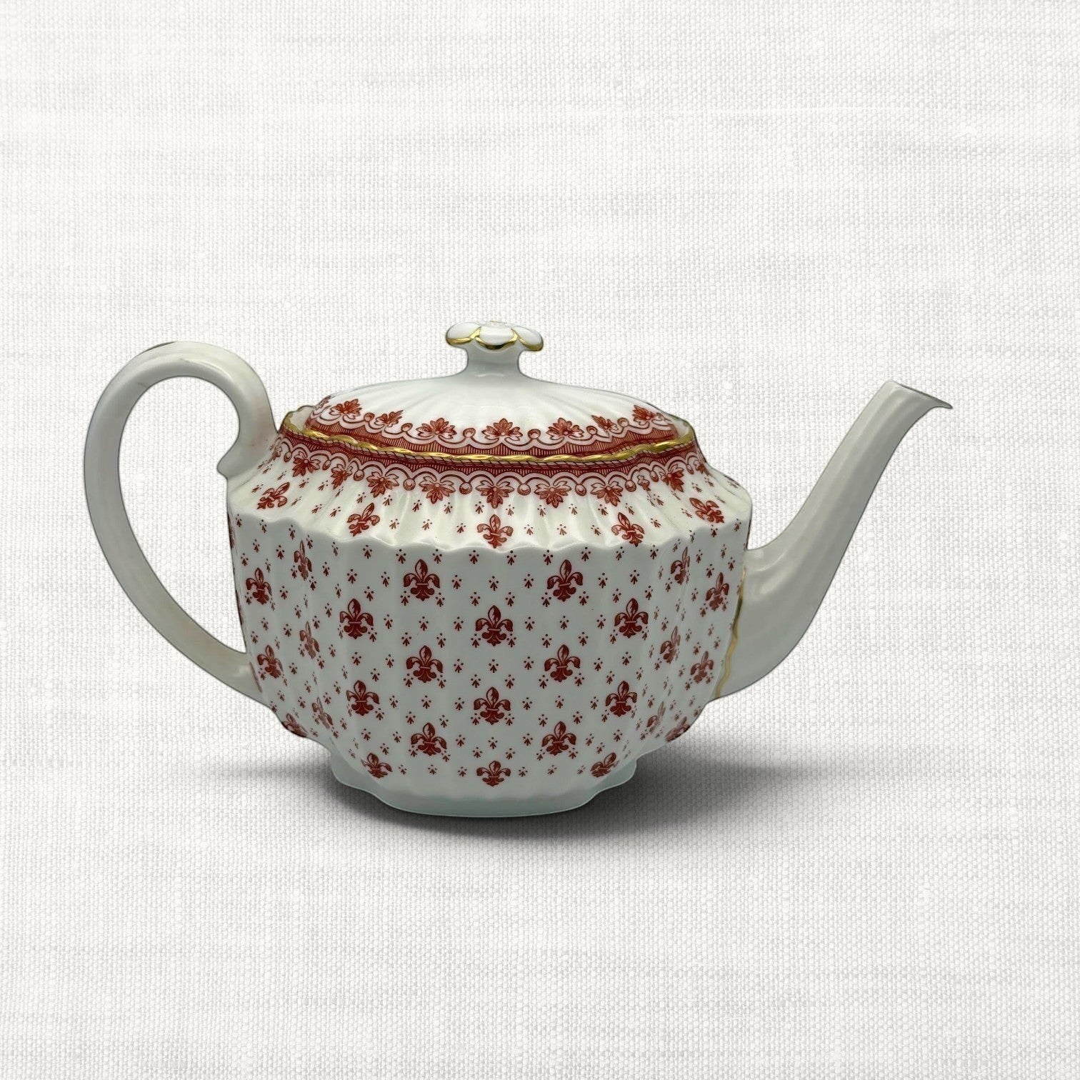 A white porcelain teapot with a repeating red floral pattern and gold trim, set against a light beige textured fabric background.