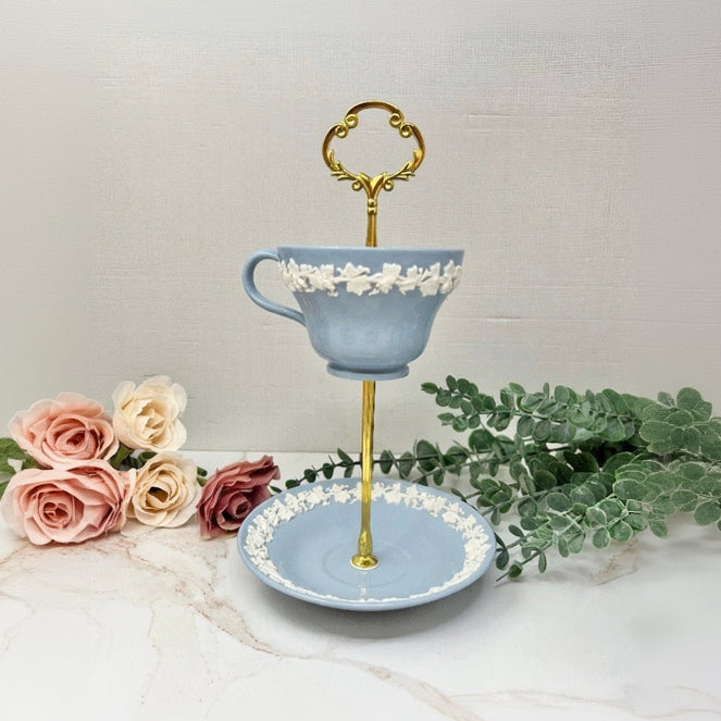 Decorative teacup stanyd ft. Wedgwood Queensware pattern with a blue teacup and gold accents, surrounded by flowers and greenery.