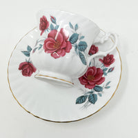 Deep pink/ruby red roses with green leaves on white bone china teacup and saucer. View of teacup resting on saucer.